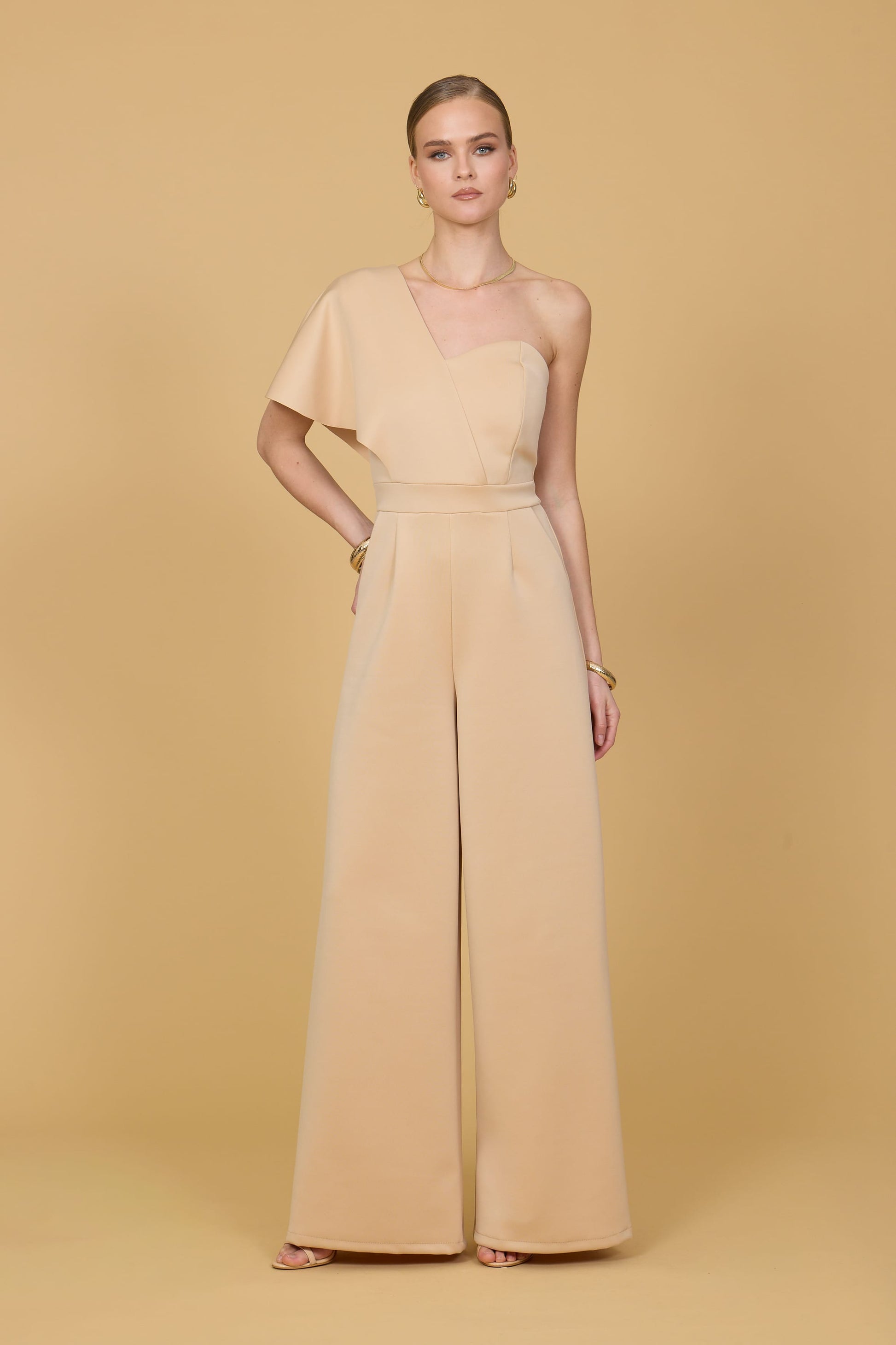 Woman wearing a light mustard one-shoulder jumpsuit on a beige background
