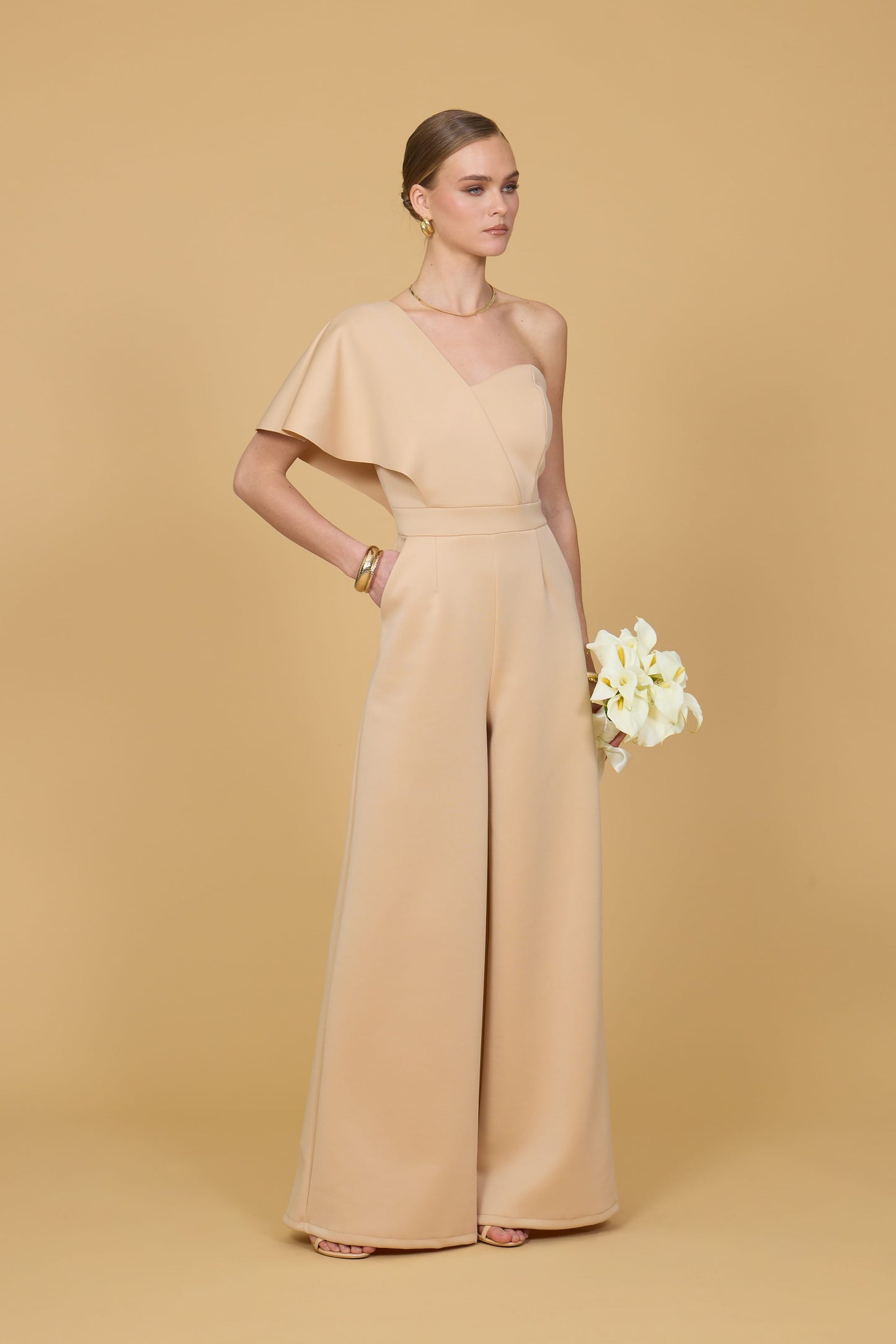 Woman wearing a light mustard one-shoulder jumpsuit on a beige background