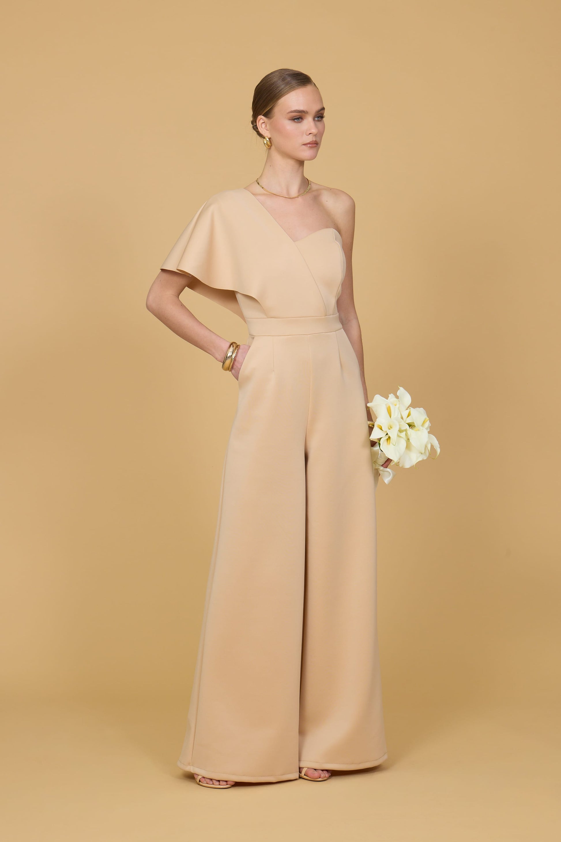 Woman wearing a light mustard one-shoulder jumpsuit on a beige background