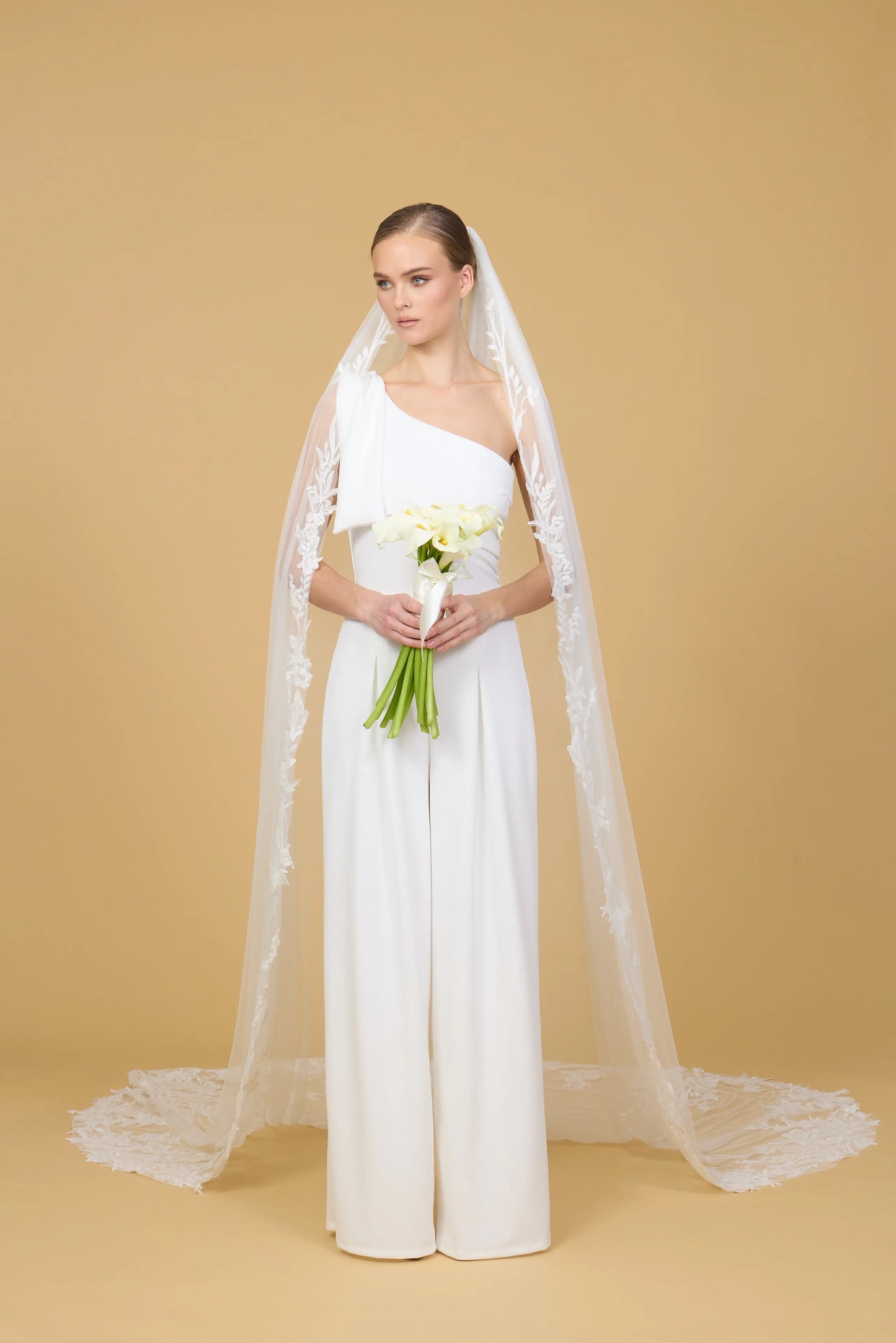 Woman in a white bridal gown with a long veil holding flowers against a beige background
