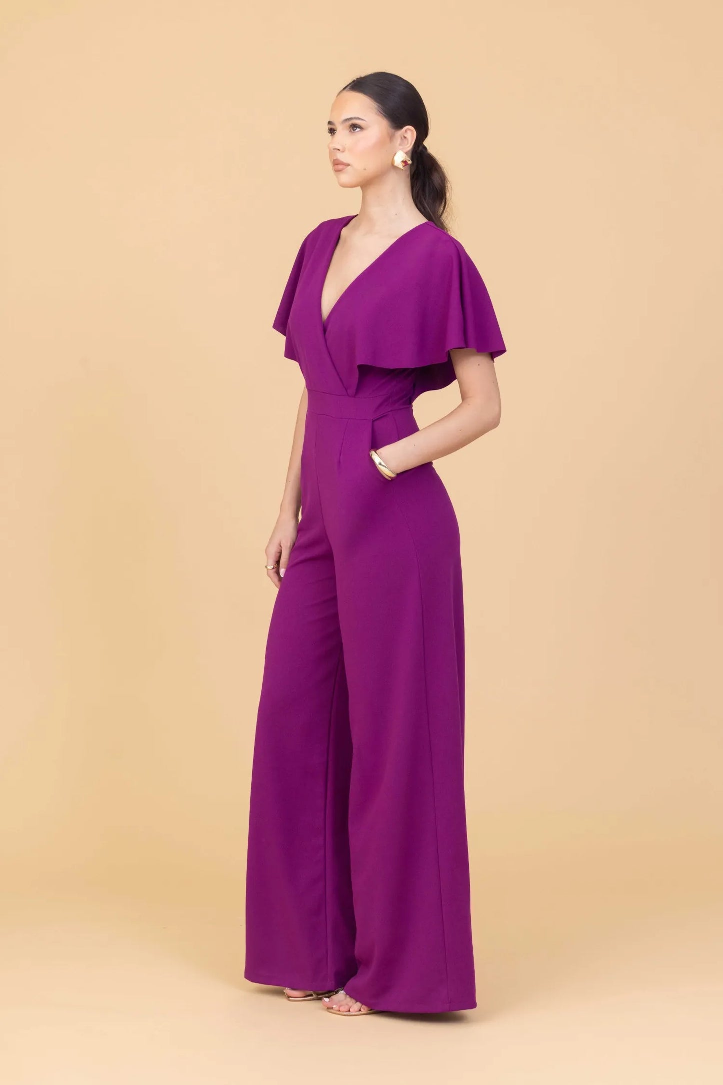 Woman wearing a purple jumpsuit with cape sleeves on a beige background