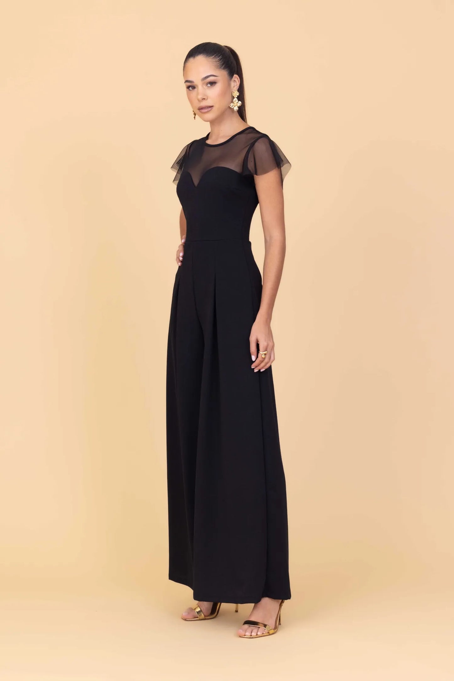 Woman wearing a black jumpsuit with sheer sleeves against a beige background